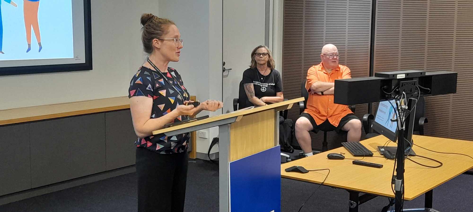image of woman in foreground at podium with two people sitting behind her