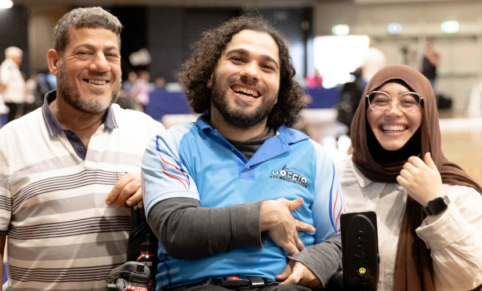 two men and a woman wearing a hijab on a sports court smiling. the man in the middle is in a wheelchair
