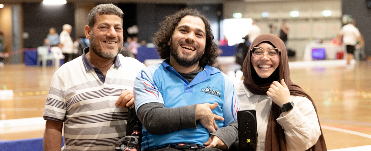 two men and a woman wearing a hijab on a sports court smiling. the man in the middle is in a wheelchair